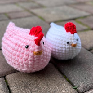 May include: Two crocheted chicken plushies, one pink with a red comb and the other white with a red comb.