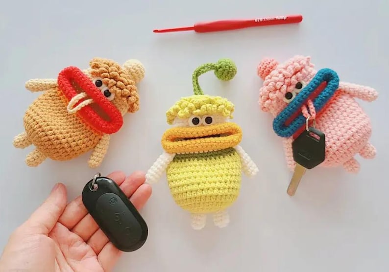 May include: Three crocheted keychains in the shape of cartoon characters. The first keychain is brown with a red mouth and a yellow nose. The second keychain is yellow with a green stem and a yellow mouth. The third keychain is pink with a blue mouth and a pink hat. Each keychain has a black key fob attached.