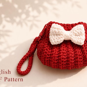 Può includere: Una pochette all'uncinetto rosso scuro con un design a coste testurizzato e un fiocco bianco all'uncinetto fissato sul davanti. La pochette include un cinturino da polso all'uncinetto rosso abbinato con una chiusura in plastica rossa. Il testo English PDF Pattern è visibile.