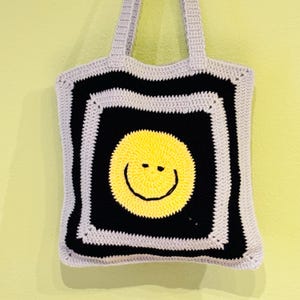Peut inclure: Sac fourre-tout crocheté avec un motif de smiley. Le sac présente un carré noir avec un smiley jaune au centre, bordé d'une bordure noire et d'une bordure grise. Les anses et le reste du sac sont gris.