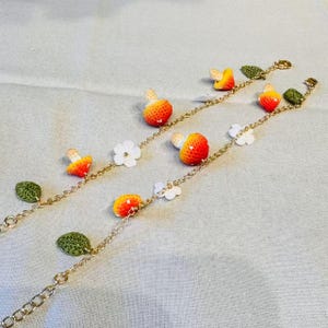 May include: Two gold-colored chain bracelets adorned with crocheted mushrooms, white flowers, and green leaves. The mushrooms are orange and red with white stems. The leaves are green, and the flowers are white. The bracelets are on a light gray surface.