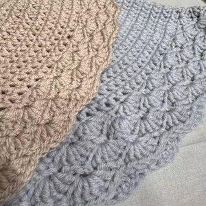 May include: Close-up of two crocheted blankets. One is a light beige color, and the other is a light gray. Both blankets have a textured, scalloped edge and a repeating pattern of raised stitches, creating a cozy, handmade appearance.