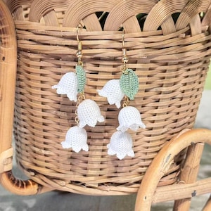 May include: White crochet lily of the valley earrings with gold-colored hooks and green crochet leaves. The earrings are displayed in front of a woven basket, showcasing a delicate floral design. The earrings are a handmade accessory.