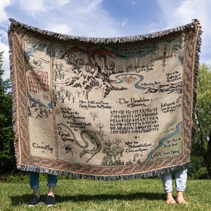 May include: A beige woven throw blanket with a map design, featuring mountains, rivers, and text. The blanket has a brown border and fringe. The blanket is held up in front of a blue sky and green trees.
