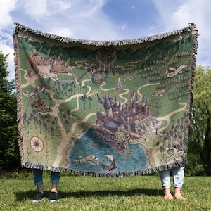 Peut inclure: Une couverture en tapisserie tissée avec un motif de carte détaillé, comprenant un château, des cours d'eau et des routes. La couverture a un code couleur vert et marron avec des bords frangés. La couverture est tenue dans un champ herbeux sous un ciel bleu.