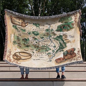 Puede incluir: Una manta beige con un mapa de La Comarca de El Señor de los Anillos. El mapa incluye características geográficas, texto e ilustraciones. La manta tiene un borde con flecos y está sostenida por dos personas.