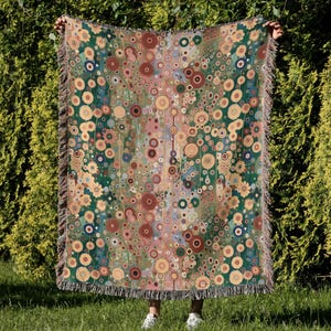 Può includere: Una coperta decorativa con un motivo ripetuto di cerchi colorati ed elementi floreali nei toni del verde, marrone e giallo. La coperta ha un bordo con frange ed è mostrata su uno sfondo di fogliame verde.