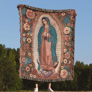 May include: Woven tapestry blanket featuring an image of the Virgin Mary surrounded by floral designs in shades of pink, blue, and brown. The blanket has a fringed edge and is held up against a blue sky background.