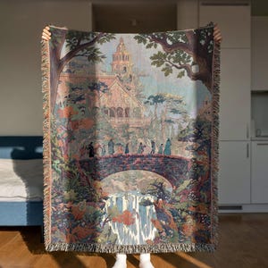 Può includere: Una coperta arazzo tessuta con una scena paesaggistica dettagliata con un ponte, una cascata e un edificio con una torre. La coperta ha un bordo a frange e una tavolozza di colori di verdi, blu e marroni.