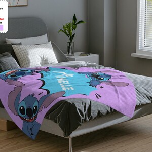 Op de afbeelding: Een lavendelkleurig pluche deken met een cartoon Stitch-personage uit de Disney-film Lilo & Stitch. Het deken heeft de tekst "Ohana betekent familie" in een blauwe spraakballon.