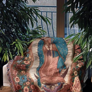 May include: A throw blanket featuring an image of Our Lady of Guadalupe. The blanket has a floral border with a fringed edge. The central image is of the Virgin Mary, with a blue cloak and a halo of stars. The color palette includes shades of brown, blue, and pink.