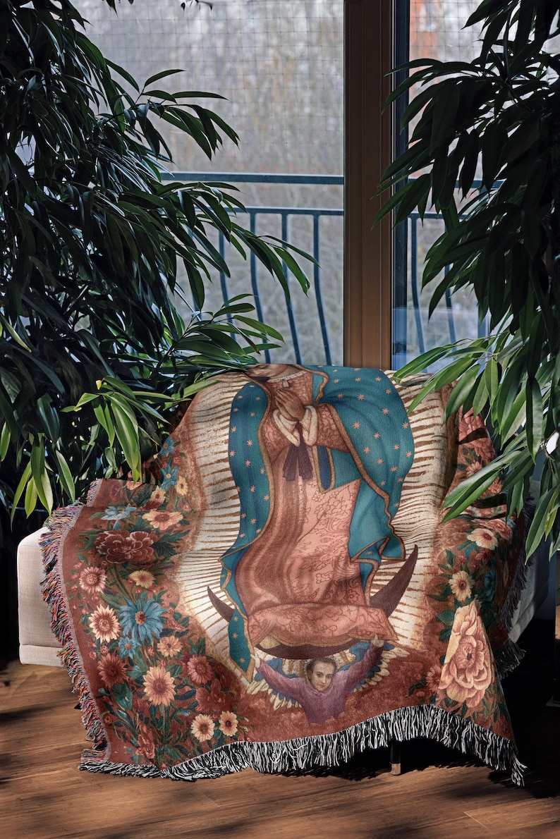 May include: A throw blanket featuring an image of Our Lady of Guadalupe. The blanket has a floral border with a fringed edge. The central image is of the Virgin Mary, with a blue cloak and a halo of stars. The color palette includes shades of brown, blue, and pink.