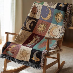 May include: A woven throw blanket draped over a wooden rocking chair. The blanket features a patchwork design with celestial imagery, including suns, moons, and stars in various colors. The blanket has a fringed edge.