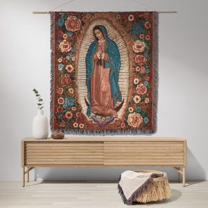 May include: A woven tapestry depicting Our Lady of Guadalupe, framed by a floral border in shades of red, orange, and blue. The central figure is draped in a blue cloak and peach-colored robes. The tapestry is hung on a wooden rod.