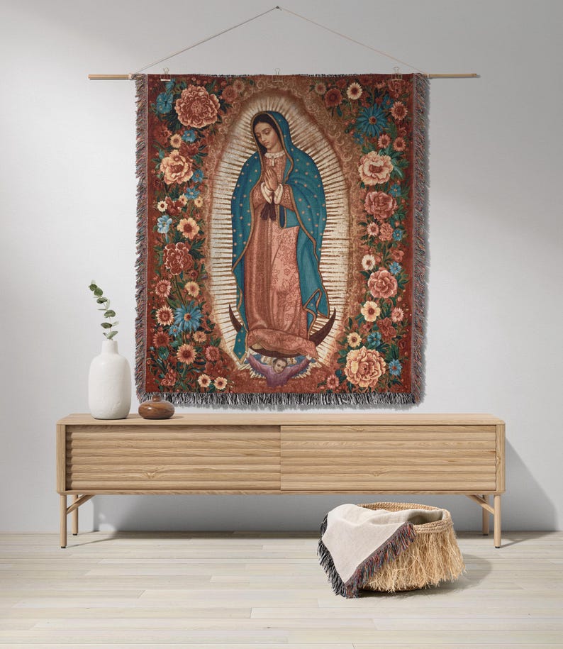 May include: A woven tapestry depicting Our Lady of Guadalupe, framed by a floral border in shades of red, orange, and blue. The central figure is draped in a blue cloak and peach-colored robes. The tapestry is hung on a wooden rod.