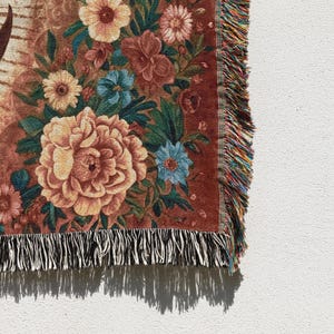 May include: A woven throw blanket with a floral design. The blanket features large peach-colored peonies, blue and yellow flowers, and green leaves against a reddish-brown background. The edge has a fringe of black and white threads.
