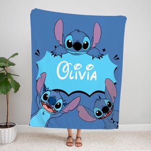 Op de afbeelding: Een blauwe fleece deken met een cartoon Stitch-personage uit de Disney-film Lilo & Stitch. Het Stitch-personage houdt een blauwe spraakballon vast met de naam "Olivia" in wit.