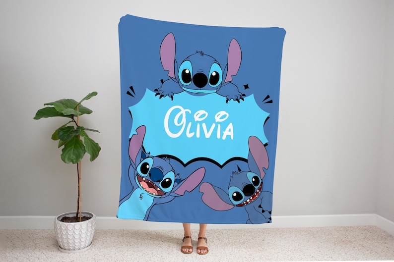 Op de afbeelding: Een blauwe fleece deken met een cartoon Stitch-personage uit de Disney-film Lilo & Stitch. Het Stitch-personage houdt een blauwe spraakballon vast met de naam "Olivia" in wit.