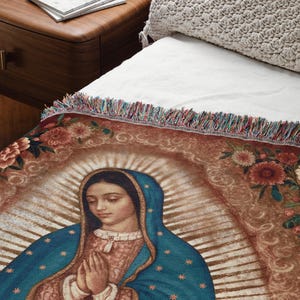 May include: A woven throw blanket featuring an image of Our Lady of Guadalupe. The blanket has a brown background with floral accents and a colorful fringe. The Virgin Mary is depicted in a blue robe with gold stars.