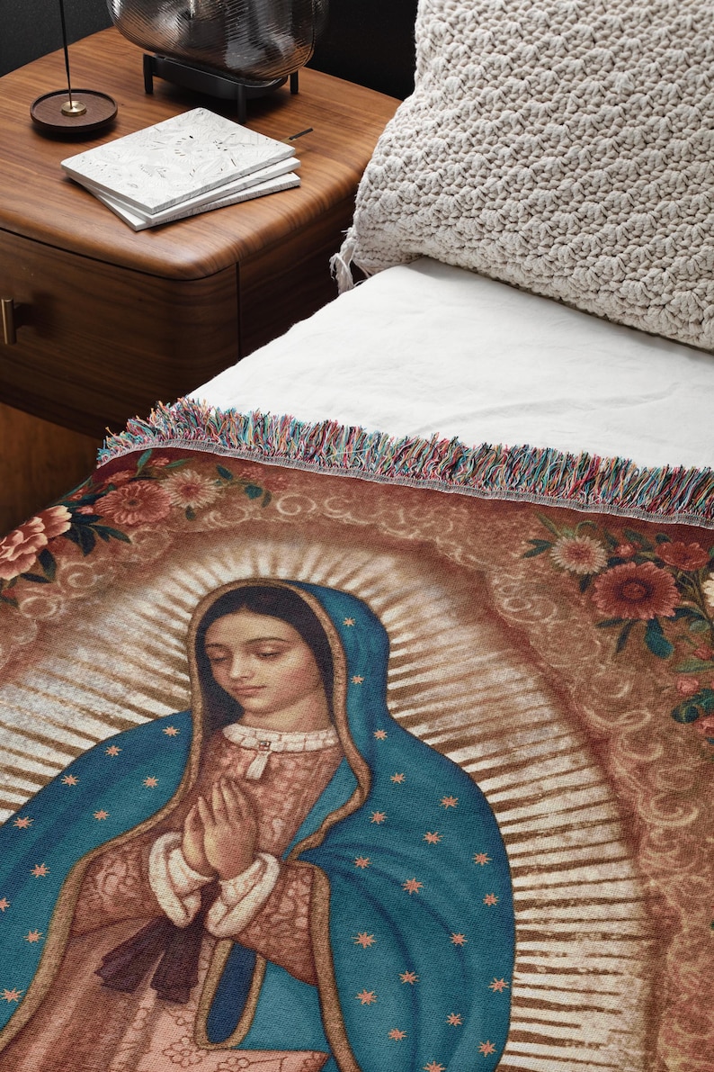 May include: A woven throw blanket featuring an image of Our Lady of Guadalupe. The blanket has a brown background with floral accents and a colorful fringe. The Virgin Mary is depicted in a blue robe with gold stars.