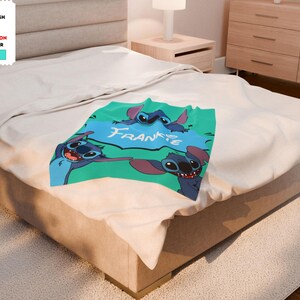 Op de afbeelding: Een turquoise blauwe, groene en witte pluche deken met drie Stitch-personages uit de Disney-film Lilo & Stitch. De deken heeft de naam Frankie in een witte spraakballon.