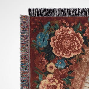 May include: A woven throw blanket featuring a floral design in shades of brown, orange, and blue. The blanket has a fringed edge with a mix of colors. The design includes large flowers and smaller floral accents.
