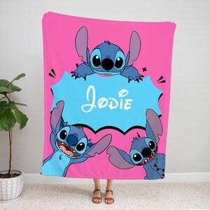 Op de afbeelding: Een roze fleece deken met een blauw cartoonfiguur, Stitch, uit de Disney-film Lilo & Stitch. De deken heeft een spraakballon met de naam "Jodie" in witte tekst.