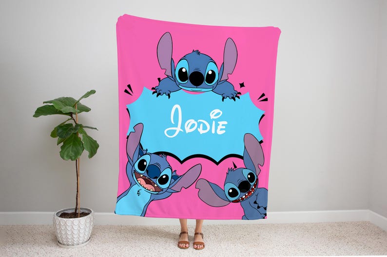 Op de afbeelding: Een roze fleece deken met een blauw cartoonfiguur, Stitch, uit de Disney-film Lilo & Stitch. De deken heeft een spraakballon met de naam "Jodie" in witte tekst.