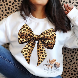 May include: White sweatshirt featuring a large leopard print bow graphic. The bow is in shades of brown and tan. A floral design and the text "Belinda Ball CREATIVE" are at the bottom right.