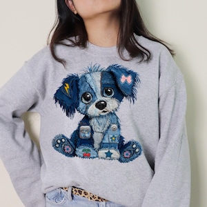 May include: A light gray sweatshirt featuring a denim-style dog graphic. The dog has blue denim patches, a pink bow, and a lightning bolt. The dog's eyes are large and expressive, and it is sitting down.