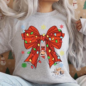 May include: A light gray sweatshirt featuring a large red bow with colorful Christmas lights and a Santa Claus figure hanging upside down. The design includes snowflakes and the text "Belinda Ball Creative".