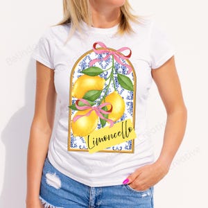 May include: White t-shirt featuring a lemon design. The design includes yellow lemons, green leaves, and pink bows, set against a blue and white patterned background. The word "Limoncello" is written in yellow.