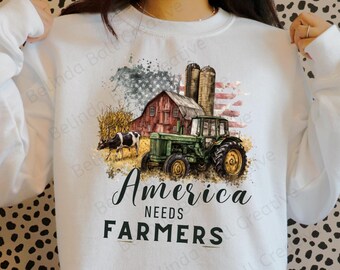Könnte beinhalten: Weißes Sweatshirt mit einer Grafik, die einen grünen Traktor, eine rote Scheune, die amerikanische Flagge und eine Kuh zeigt. Der Text "America Needs Farmers" ist in schwarzer Schrift dargestellt.