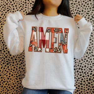 May include: White sweatshirt with the word "AMEN" embroidered across the chest. The letters are a mix of patterns, including plaid and leopard print, with orange and red accents. The sweatshirt has long sleeves.