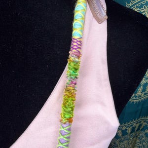 Può includere: Un'acconciatura colorata con fili blu, gialli, viola, verdi e rosa. L'accessorio è dotato di una clip metallica ed è progettato per essere attaccato ai capelli. Il design è un mix di elementi avvolti e strutturati.