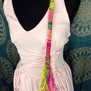 Può includere: Un'acconciatura colorata con un vivace mix di fili rosa, verdi e gialli. L'acconciatura è adornata con un fiore decorativo e una frangia di fibre luminose e strutturate. Si attacca con una piccola clip.