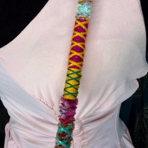 Può includere: Un'acconciatura colorata e fatta a mano con un vivace mix di trame e colori. L'accessorio è adornato principalmente con sfumature di rosa, giallo, verde acqua e marrone, creando un accessorio visivamente accattivante. Il design incorpora elementi avvolti e intrecciati.