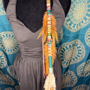 Può includere: Un'acconciatura colorata con piume arancioni, fili intrecciati e cuciture decorative. L'acconciatura è adornata con vari colori, tra cui arancione, blu e bianco, ed è fissata con una forcina nera. Questo accessorio per capelli è progettato per essere indossato nei capelli.