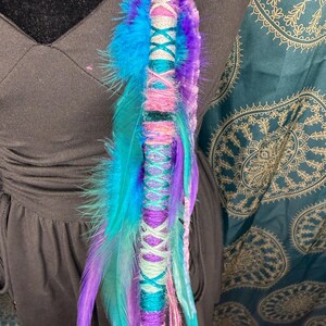 Può includere: Un'acconciatura colorata con piume blu, turchesi, viola e rosa. L'involucro è adornato con filo colorato abbinato e un nappa nella parte inferiore. Il design è accentuato con filo d'argento e un motivo a punto croce turchese e rosa.