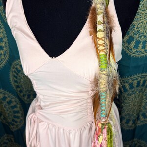 Può includere: Un abito rosa pallido con scollo all'americana, scollatura a V e dettagli arricciati. Un accessorio decorativo per capelli con piume, filo e perline è attaccato all'abito. L'accessorio per capelli presenta accenti marroni, verdi e rosa.