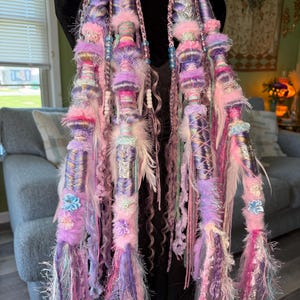 May include: Two long, decorative hair wraps in shades of pink, purple, and blue. The wraps are adorned with feathers, beads, and fabric embellishments. The wraps are designed to be worn as hair accessories, adding a whimsical touch to any hairstyle.