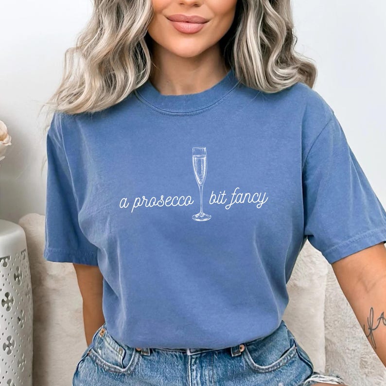May include: A blue t-shirt with a white graphic of a champagne flute and the text "a prosecco bit fancy".