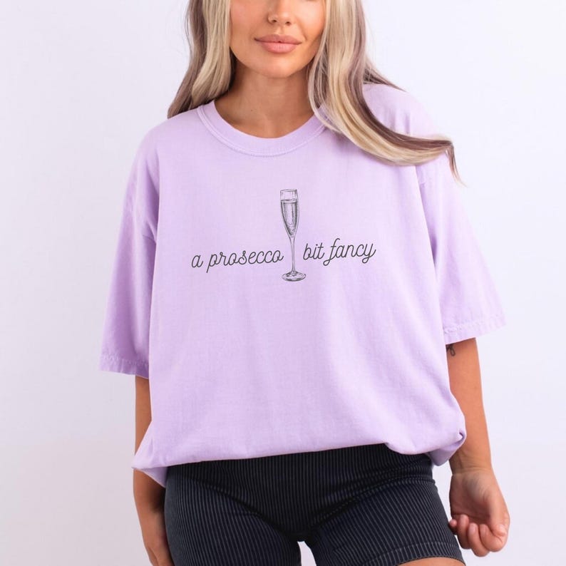 May include: A lavender t-shirt with a graphic of a champagne flute and the text "a prosecco bit fancy".