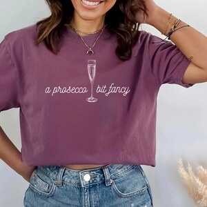 May include: A mauve t-shirt with a white graphic of a champagne flute and the text "a prosecco bit fancy".