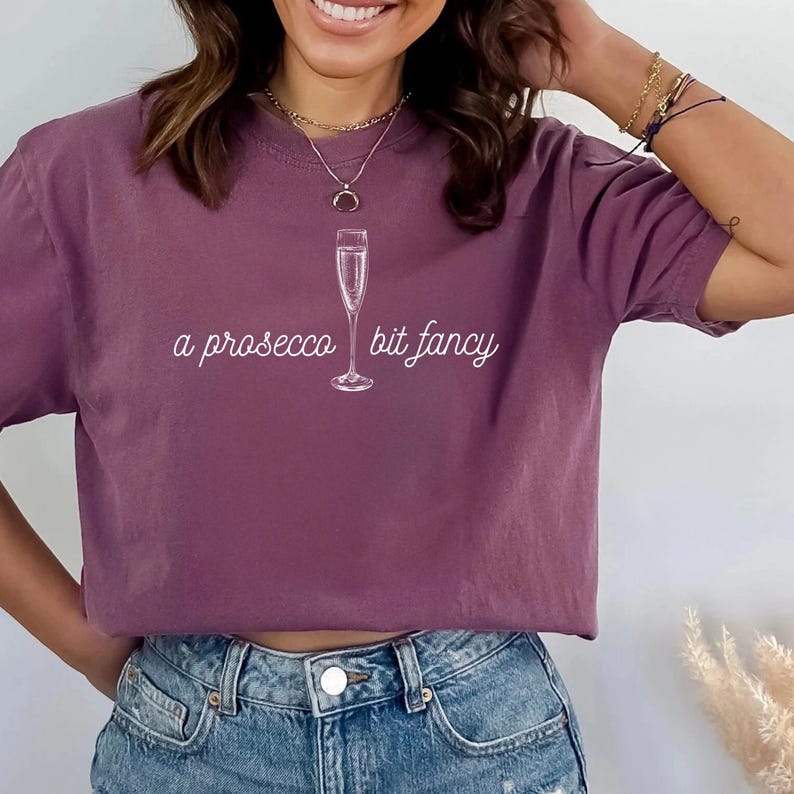 May include: A mauve t-shirt with a white graphic of a champagne flute and the text "a prosecco bit fancy".