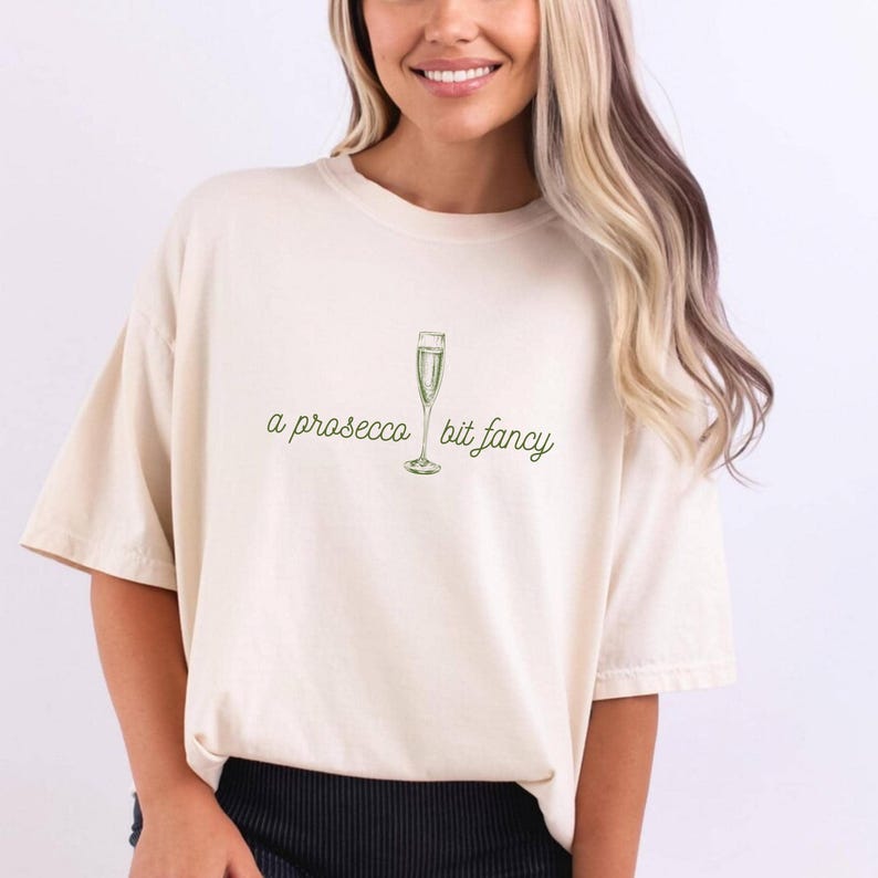 May include: A white t-shirt with a green illustration of a champagne flute and the text "a prosecco bit fancy".