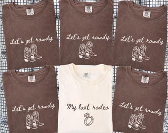 May include: Six t-shirts, some espresso-colored and some ivory-colored, are displayed. The espresso shirts read "Let's get rowdy" with cowboy boot graphics. The ivory shirt says "My last rodeo" with a ring graphic.