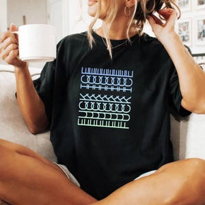 May include: Black t-shirt with a gradient blue and green graphic design. The design features several rows of different stitch patterns. The shirt is a comfortable, casual style.