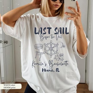May include: White t-shirt with a navy blue graphic that reads "LAST SAIL Before the Veil" and "Kamila's Bachelorette Miami, FL". The design includes a ship's wheel, champagne glass, ring, and seashells. The shirt has a denim print.