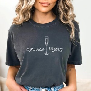 May include: A dark gray t-shirt with a white graphic of a champagne flute and the text "a prosecco bit fancy".